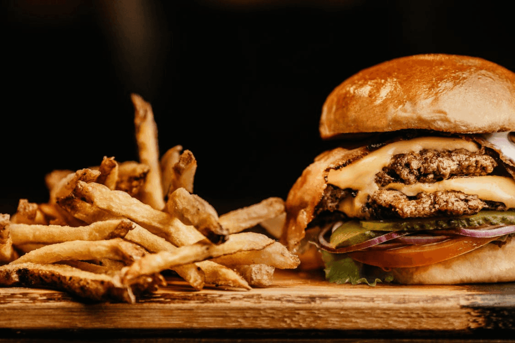 A close-up of crispy French fries next to a double cheeseburger with lettuce, tomato, red onion, pickles, melted cheese, and a shiny brioche bun on a wooden surface.