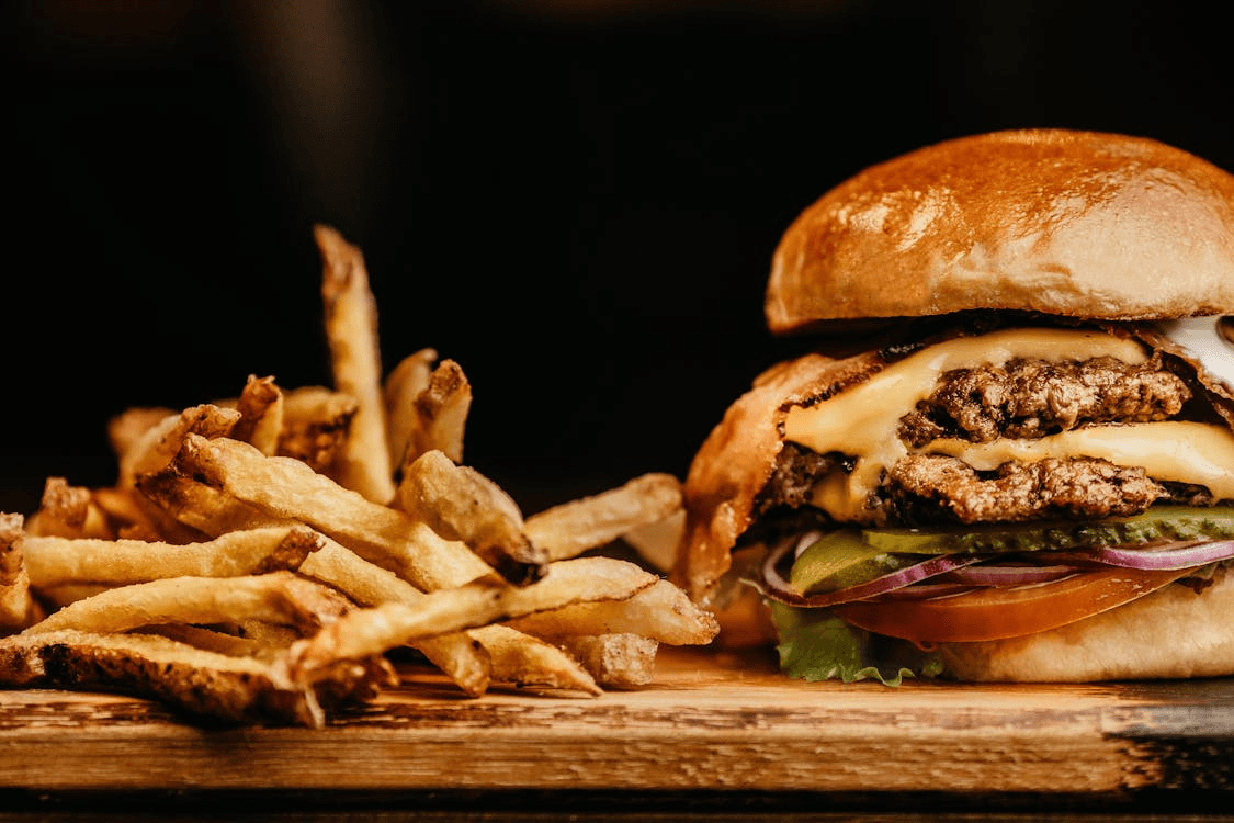 A close-up of crispy French fries next to a double cheeseburger with lettuce, tomato, red onion, pickles, melted cheese, and a shiny brioche bun on a wooden surface.
