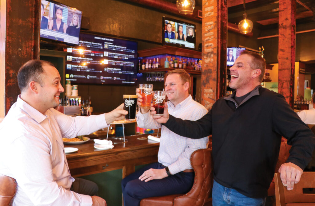Three men sit at a bar, smiling and laughing as they toast with beer glasses. TV screens and drinks are visible in the background, creating a lively, casual atmosphere.