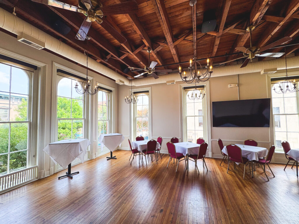 A bright event room with large arched windows, wooden floors, exposed wooden ceiling beams, chandeliers, and several round and square tables with white tablecloths and red chairs. A large flat-screen TV is mounted on the wall.
