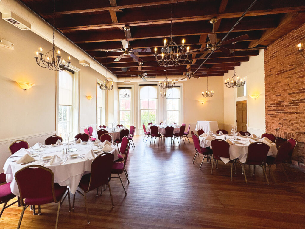 A banquet hall with round tables covered in white cloths and burgundy chairs, set for a formal event. Chandeliers hang from a wooden beamed ceiling and large windows let in natural light.