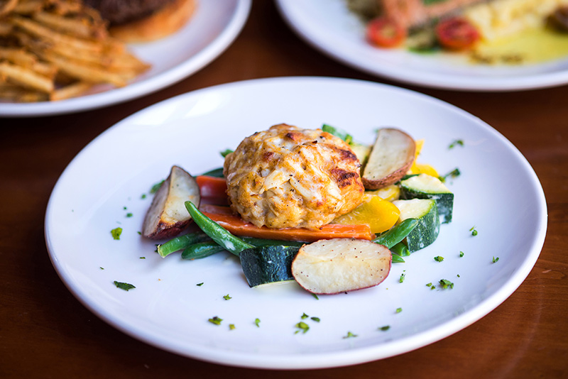 A crab cake served on a white plate with roasted potato wedges, green beans, carrots, yellow squash, and zucchini, garnished with chopped parsley.