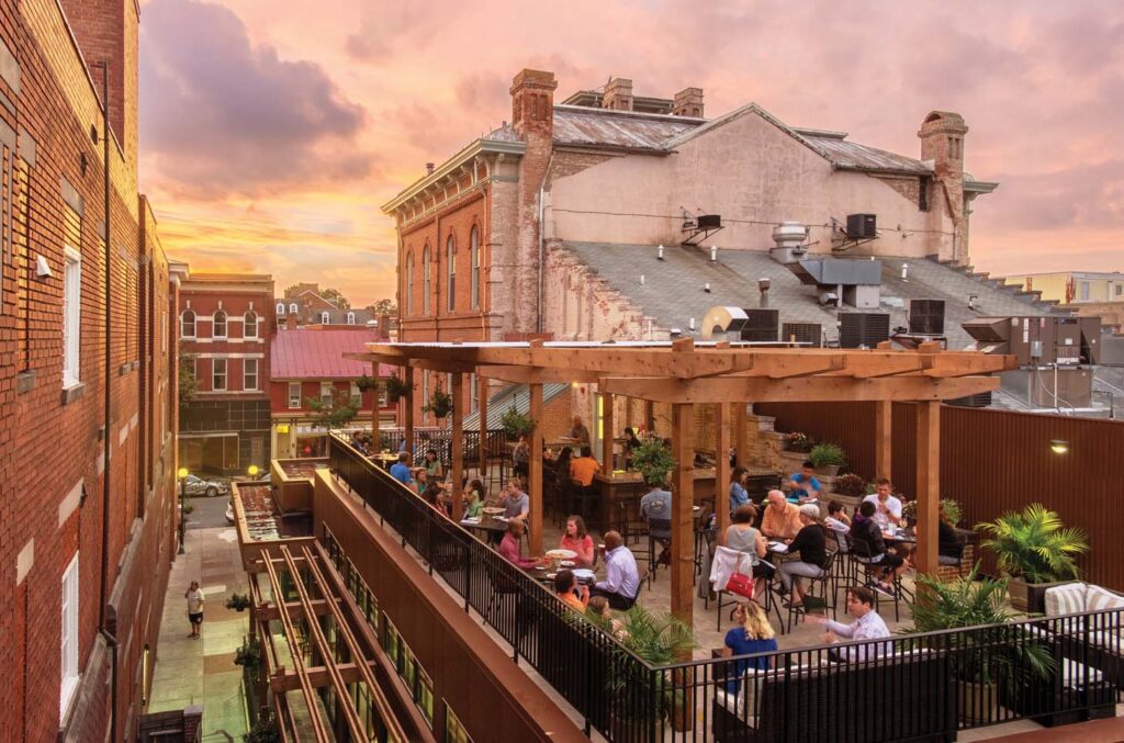 People dine and socialize on a rooftop patio with wooden pergolas and potted plants, surrounded by historic brick buildings during a colorful sunset. The scene is lively and inviting in an urban setting.