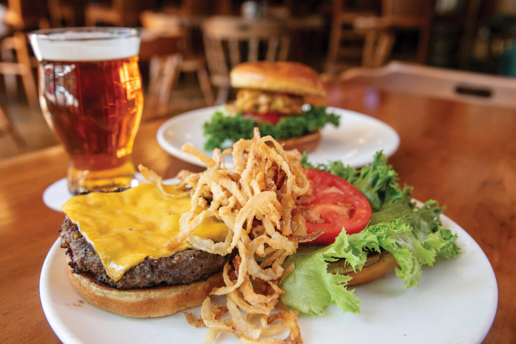 A close-up of a cheeseburger with crispy fried onions, lettuce, and tomato on a white plate, with a glass of beer and another burger in the background on a wooden table.