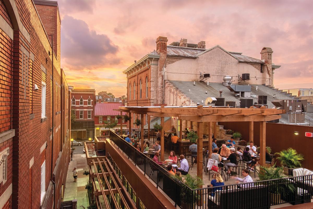 A lively rooftop restaurant with people dining under a wooden pergola at sunset, surrounded by historic brick buildings and a colorful sky.