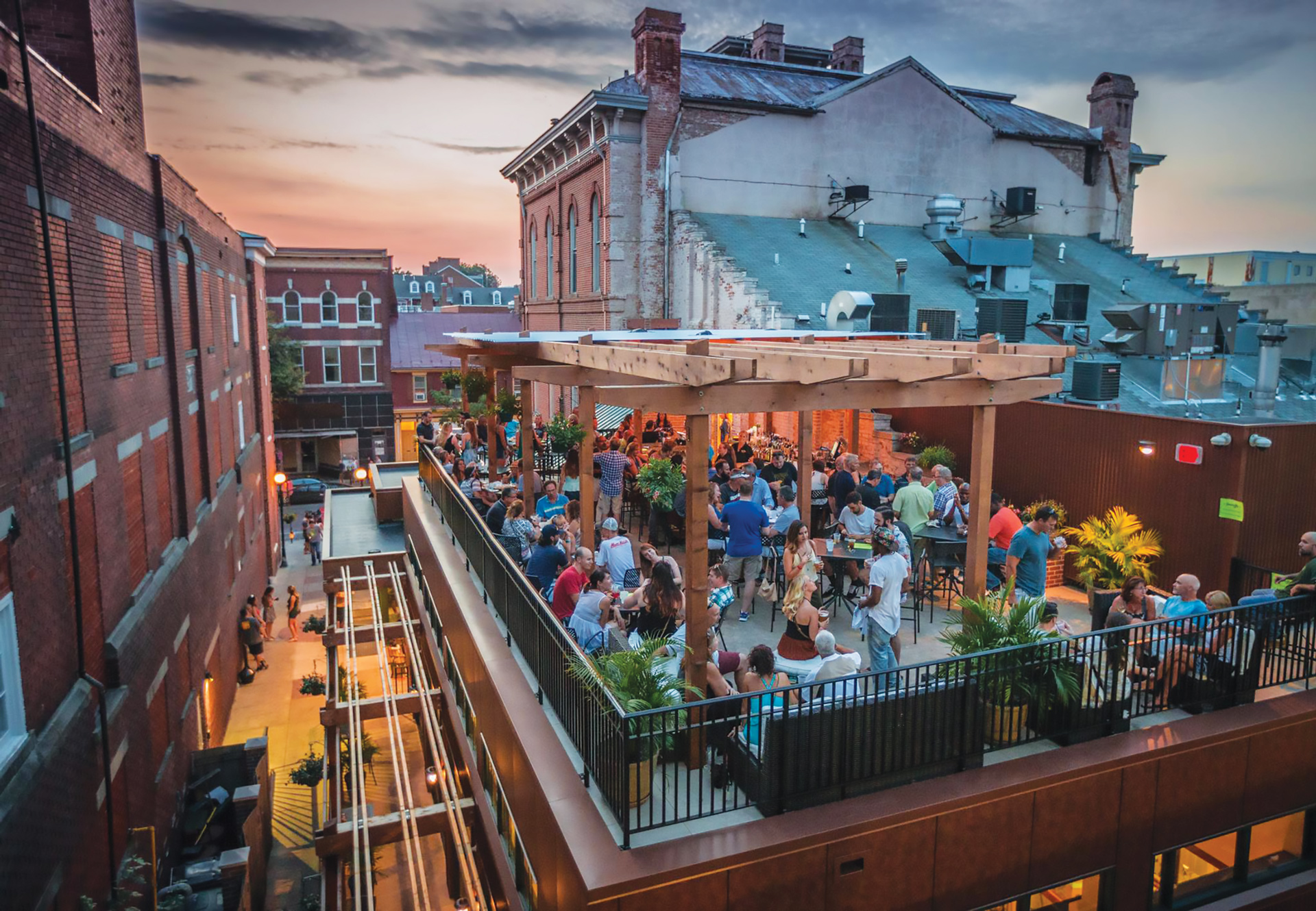 A lively rooftop bar is filled with people socializing at sunset, surrounded by brick buildings and plants, with a view of an urban alley below.