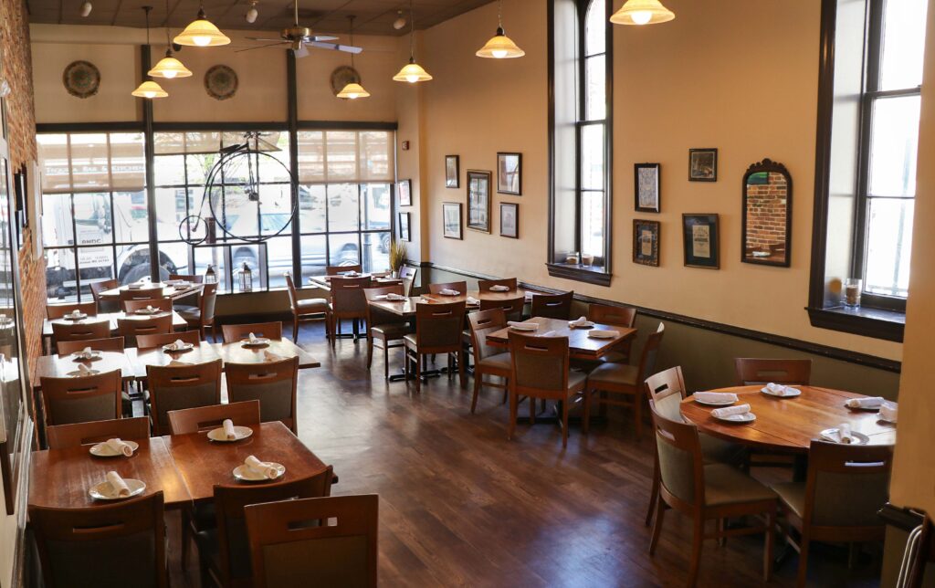 A cozy, well-lit restaurant dining area with wooden tables and chairs, place settings arranged neatly, artwork and mirrors on cream-colored walls, and large windows letting in natural light.