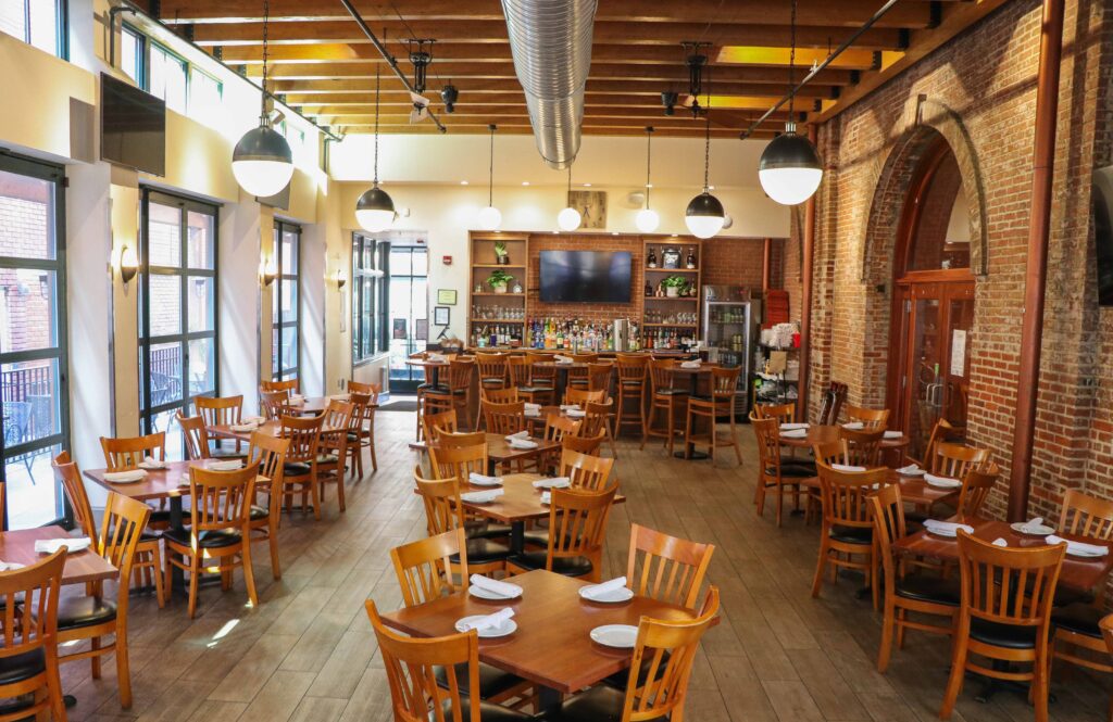Spacious restaurant with wooden tables and chairs, set with white plates. Large bar area in the back, exposed brick walls, ceiling beams, hanging lights, and large windows provide natural light.