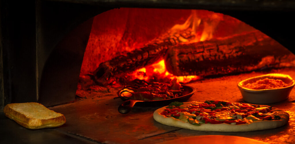 A pizza with fresh toppings, a small loaf of bread, a skillet dish, and a bowl of cheese are inside a wood-fired oven with glowing embers and flames in the background.