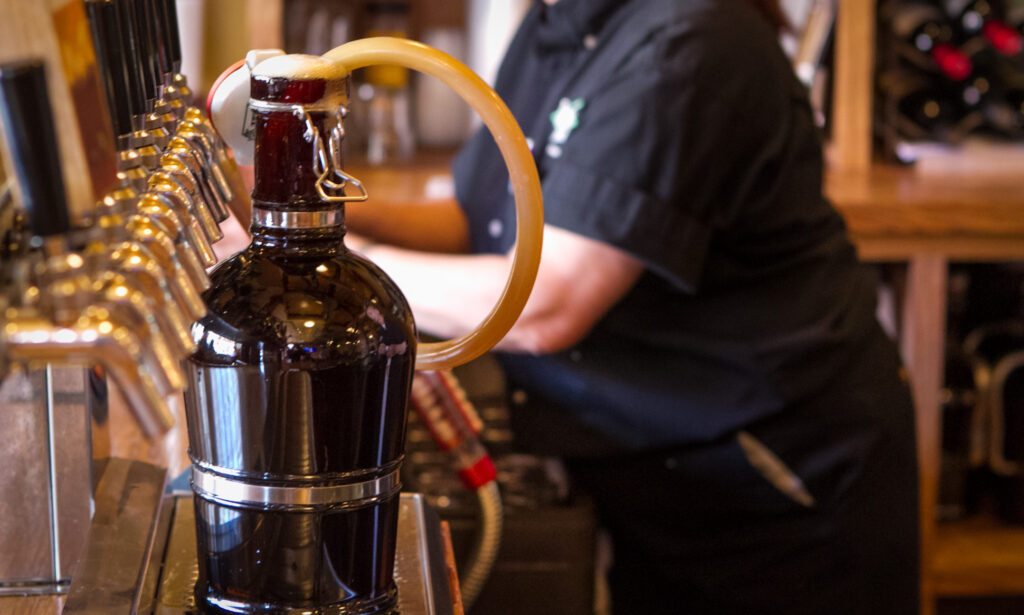 A dark growler is being filled with beer from a tap at a bar, using a yellow hose. A person in a black shirt stands in the background, slightly out of focus.