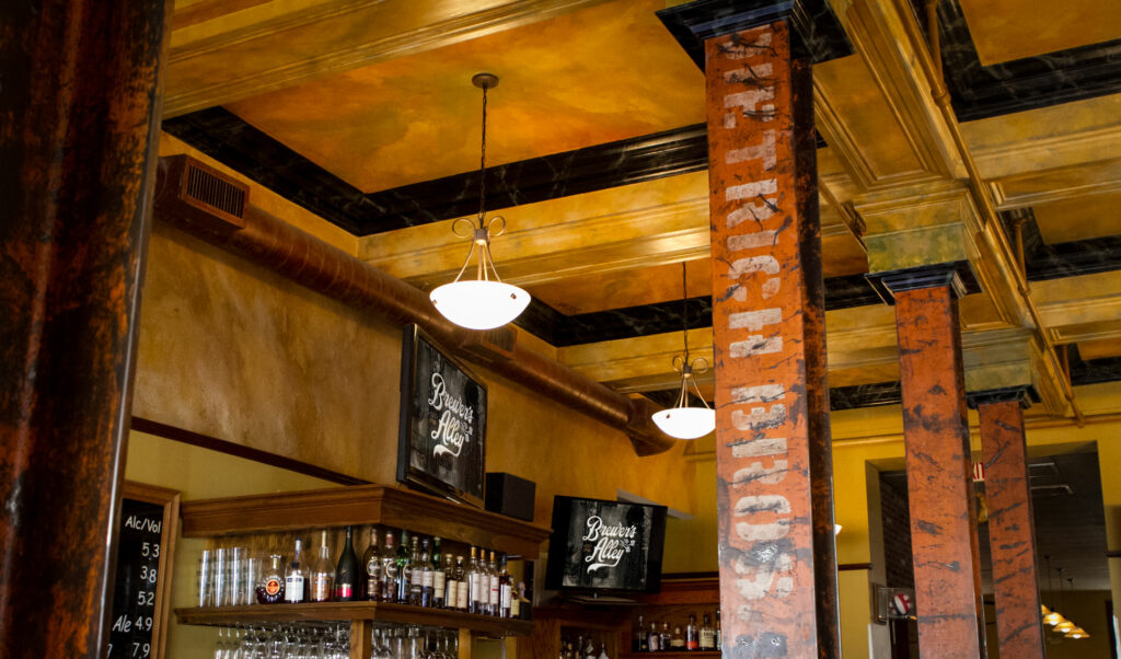 Interior of a bar with wooden shelves holding bottles, chalkboard drink menus, two pendant lights, and decorative columns with distressed paint near a gold and brown painted ceiling.
